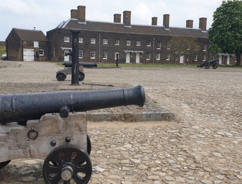 Cannon in foreground of cobblestone courtyard. A row of brown brick terrace houses are in the distance.