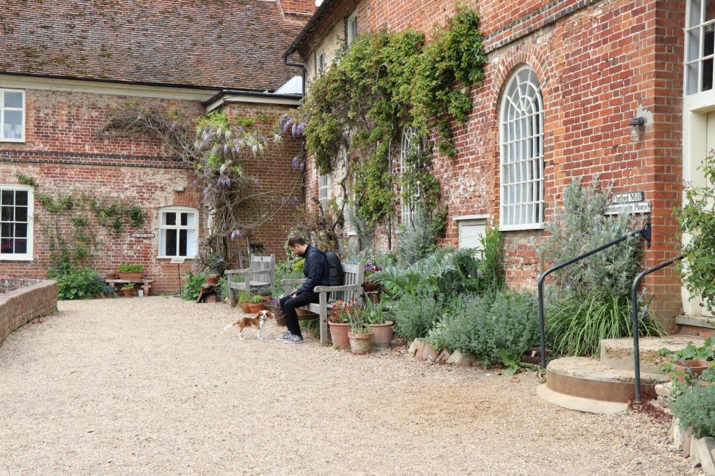 Man and dog sat on bench in front of red brick Flatford Mill building